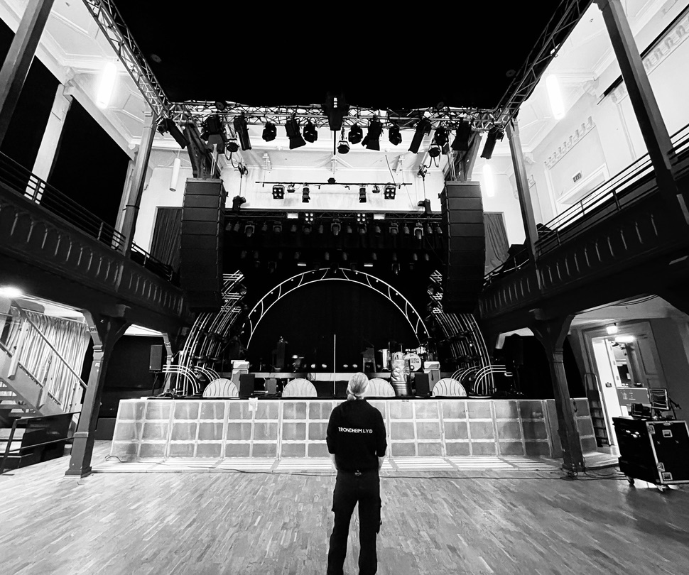 Black-and-white view inside Byscenen, with suspended PA arrays on either side of the stage and a technician on the empty floor.