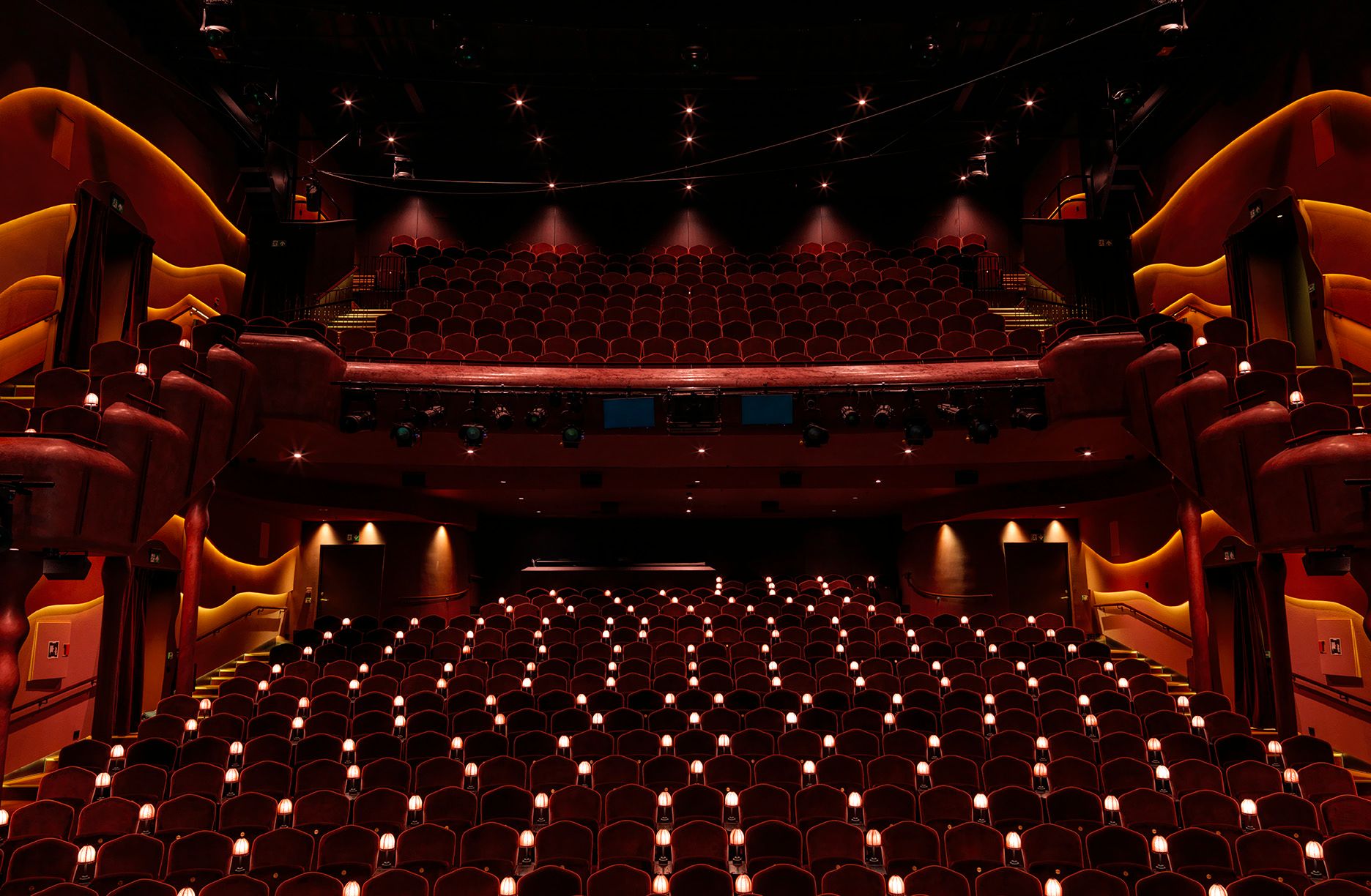 Nye Hjorten Teater’s empty auditorium with red seats, curved balconies and warm lighting, viewed from the stage.